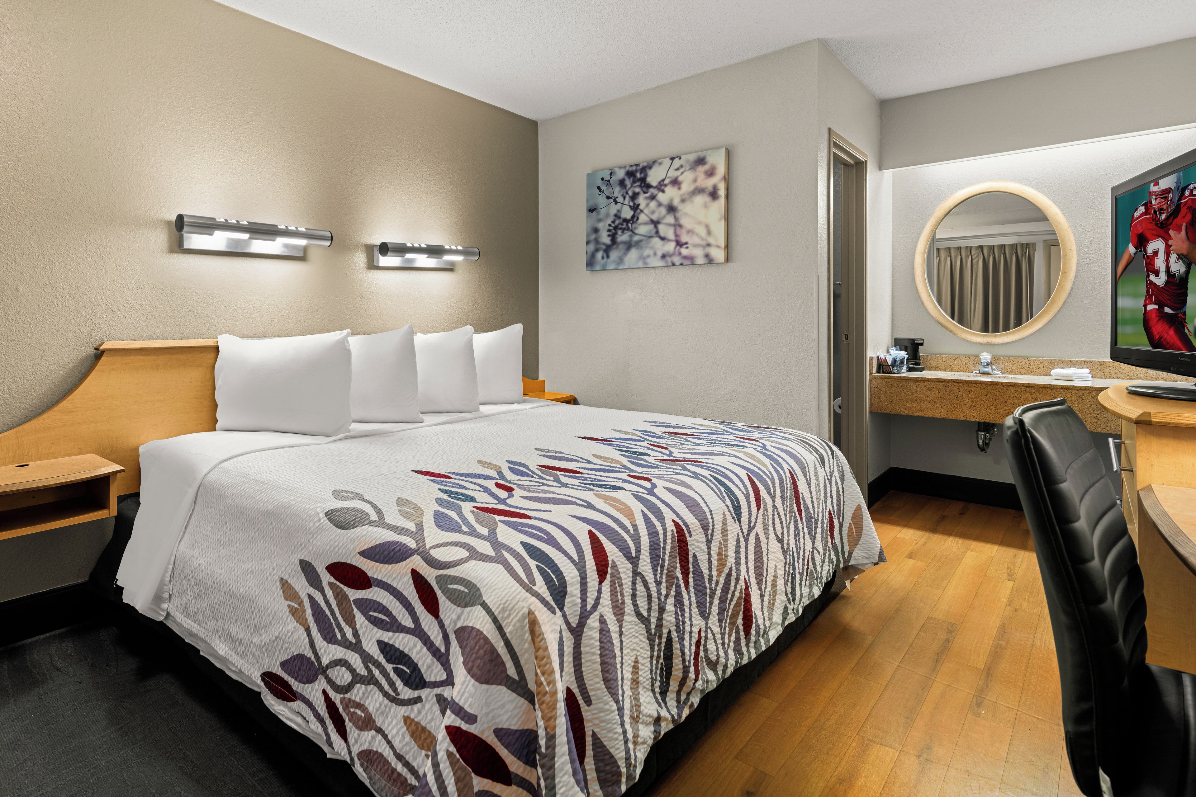 A hotel room with a bed, bedside tables, a desk with a chair, a mirror, and a wall-mounted TV.