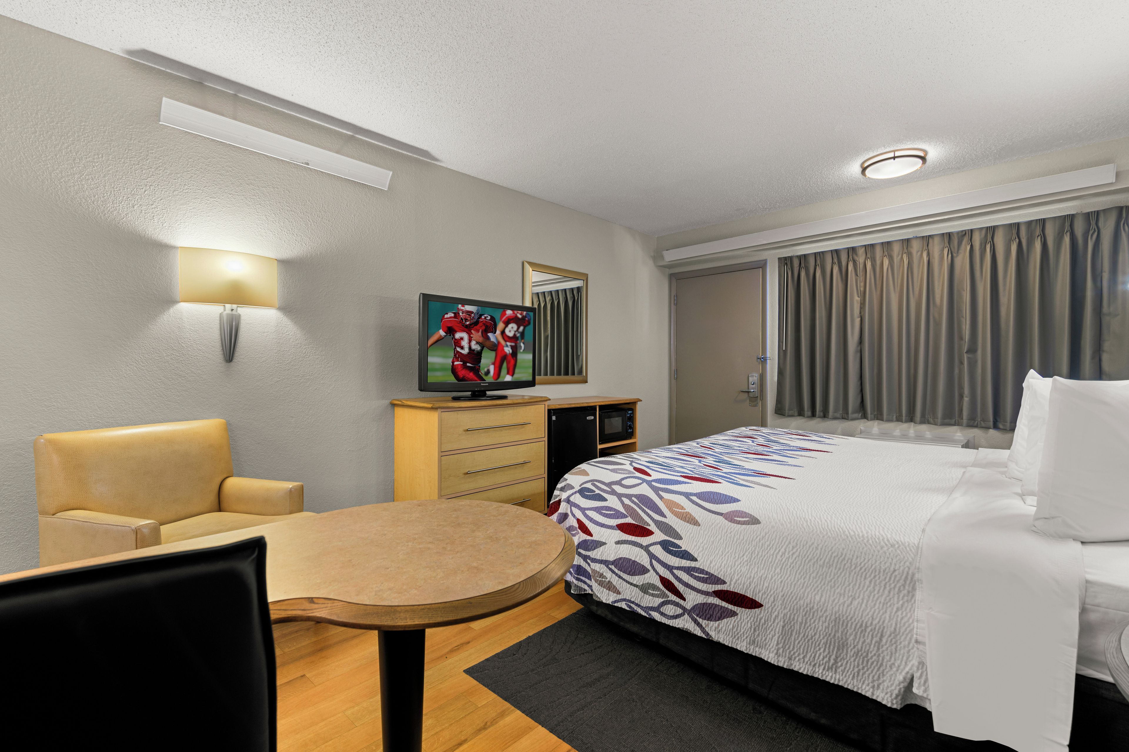 Une chambre d’hôtel équipée d’un lit, d’un bureau, d’une chaise, d’une télévision, d’un miroir et d’une fenêtre avec des rideaux.