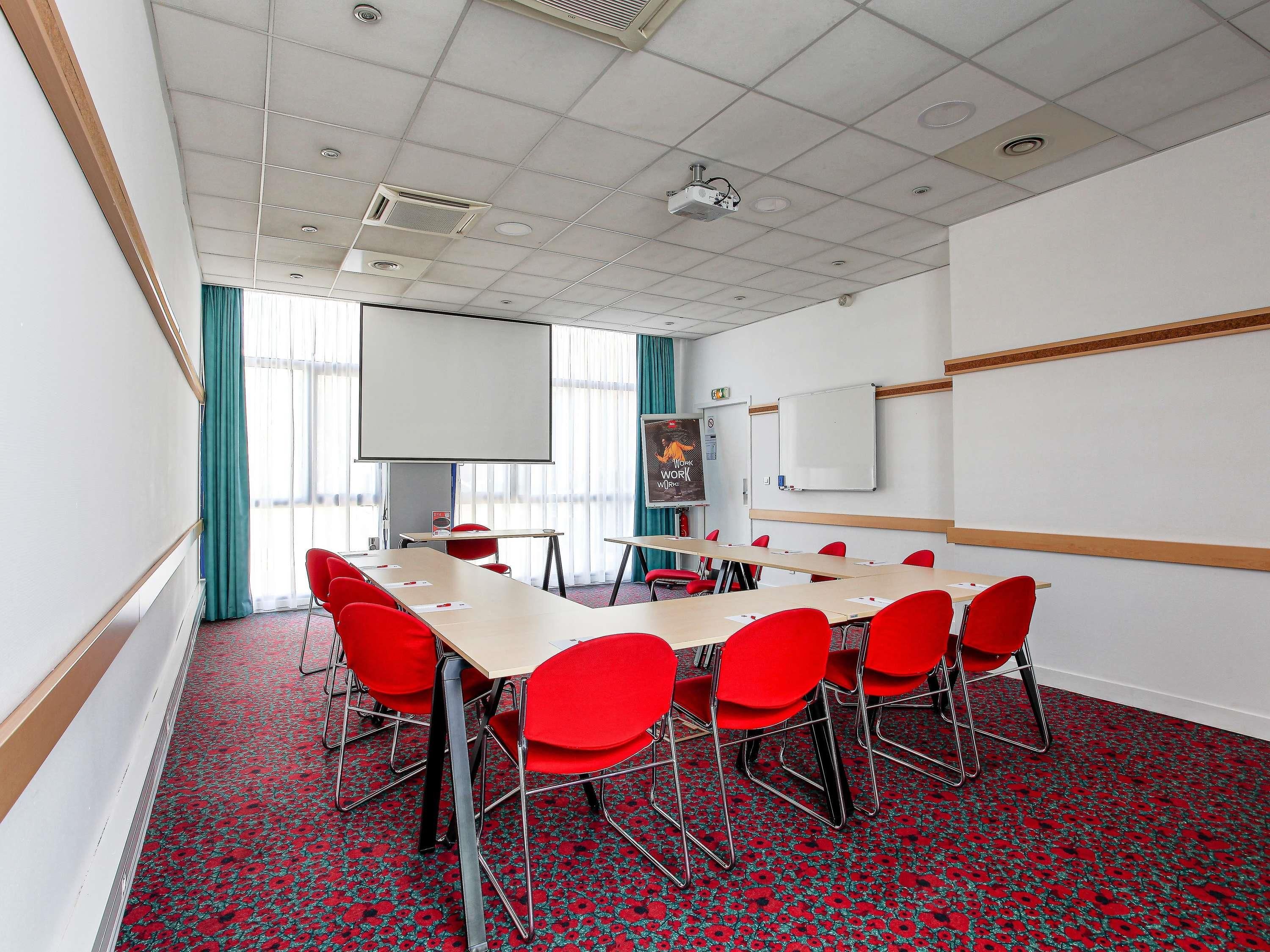 Een vergaderzaal met een grote tafel, rode stoelen, een whiteboard en een projectiescherm.