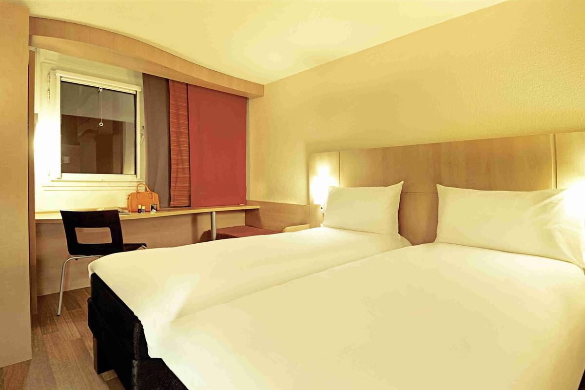 A hotel room with a double bed, a desk with a chair, and a window with curtains.