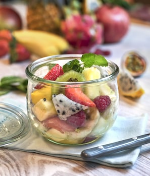 A clear glass bowl filled with assorted fruits and a mint leaf, placed on a cloth with a knife beside it.