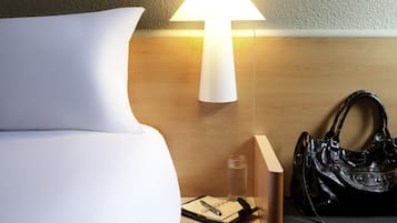 En pænt redt seng med en hvid pude, et natbord med en lampe, et glas vand, en notesbog, en pen og en sort taske.
