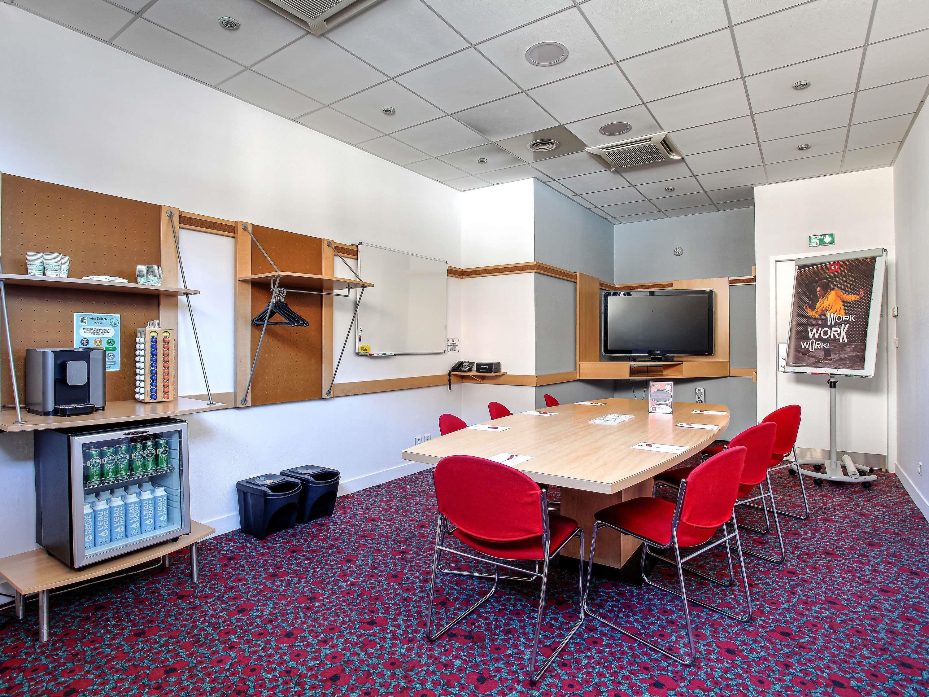 A conference room with a large table, red chairs, a whiteboard, a television, and a refrigerator with beverages.