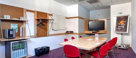 A conference room with a large table, red chairs, a whiteboard, a television, and a refrigerator with beverages.