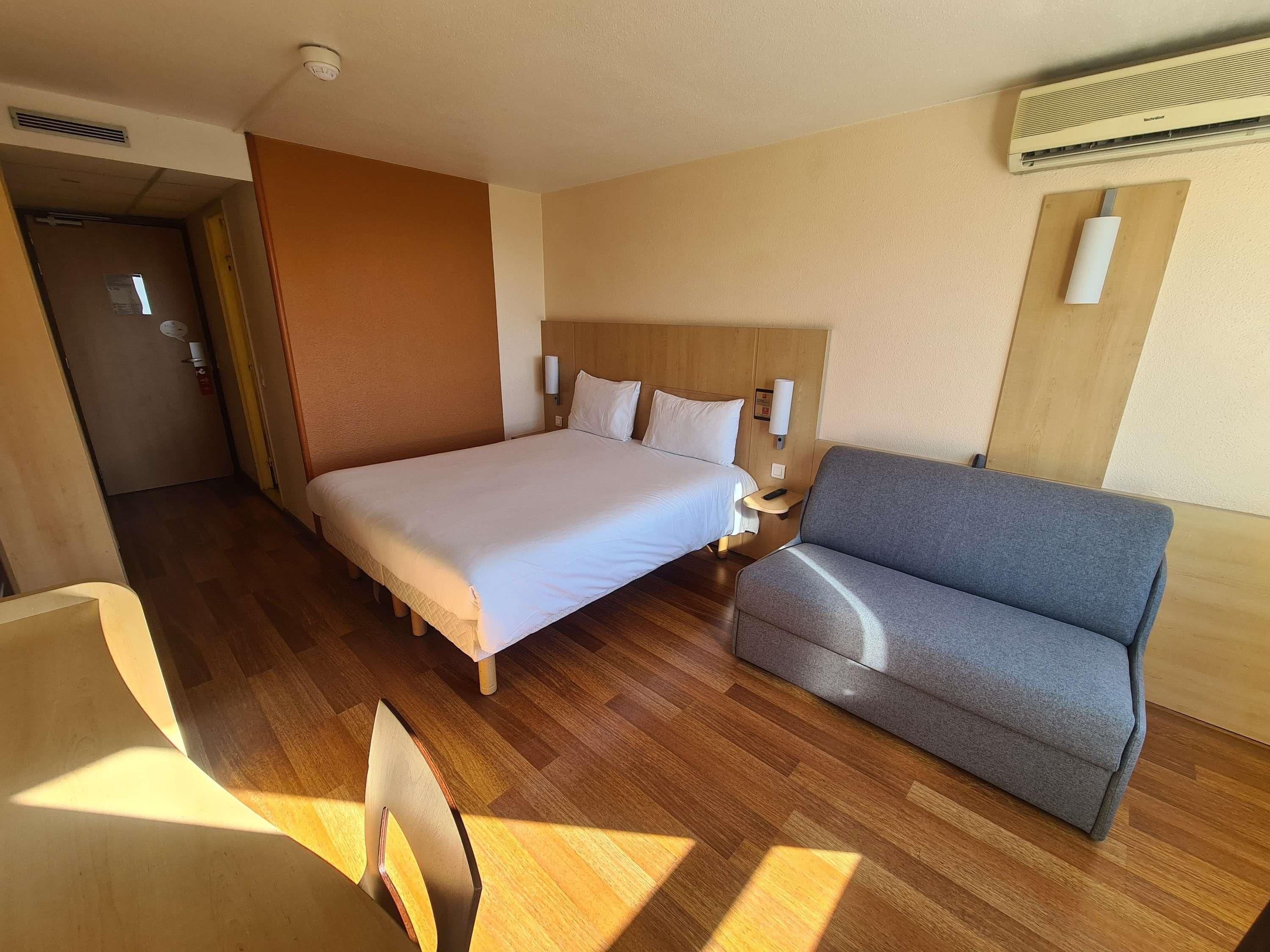Habitación de hotel con una cama grande, un sofá gris, piso de madera y aire acondicionado instalado en la pared.