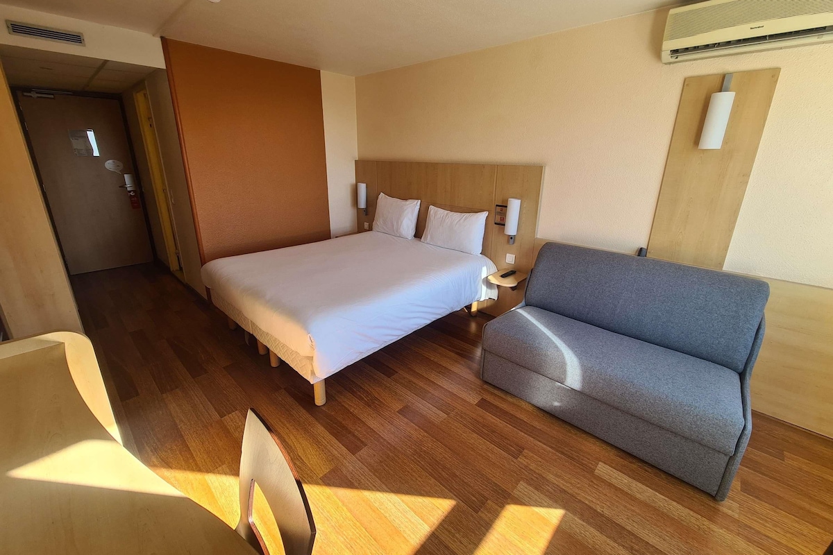 A hotel room with a large bed, a gray sofa, wooden flooring, and a wall-mounted air conditioner.