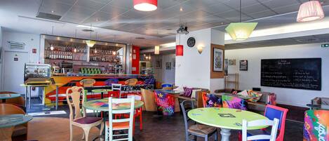 A cafe with colorful chairs and tables, a bar area with shelves of bottles, and a chalkboard with writing.