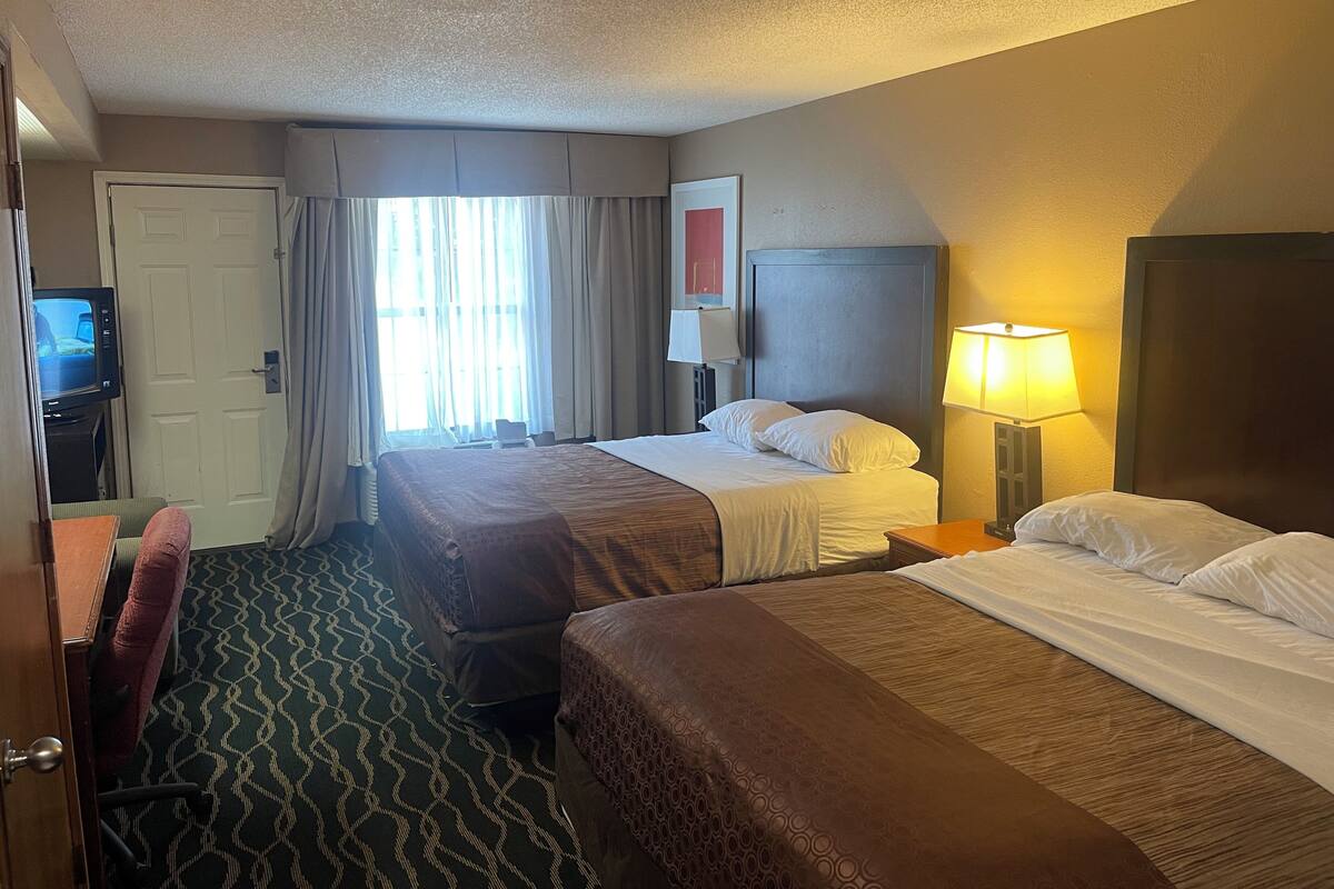 A hotel room with two beds, a desk with a television, a lamp, and a window with curtains.