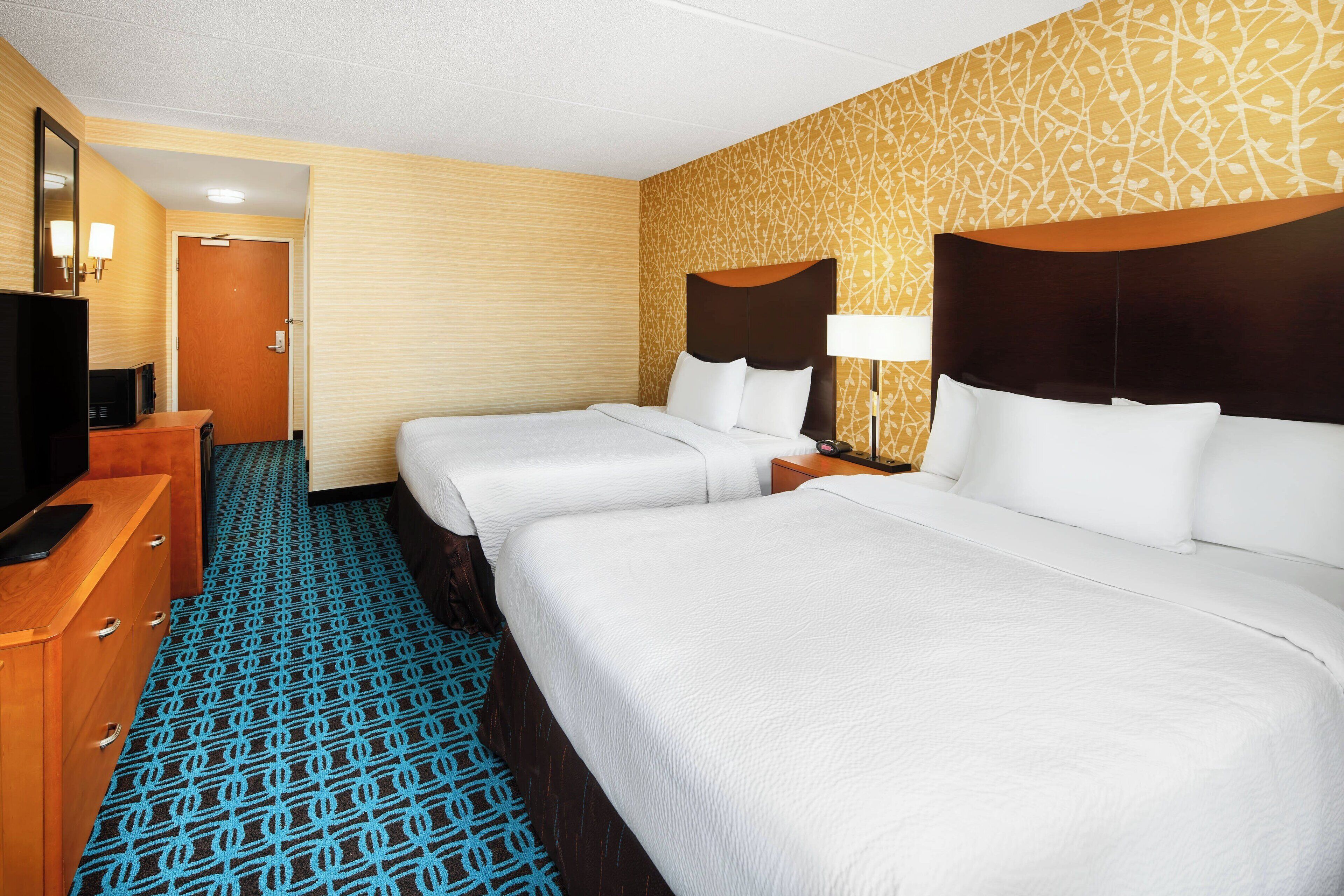 Une chambre d’hôtel avec deux lits, une commode en bois, un téléviseur et une moquette à motifs.