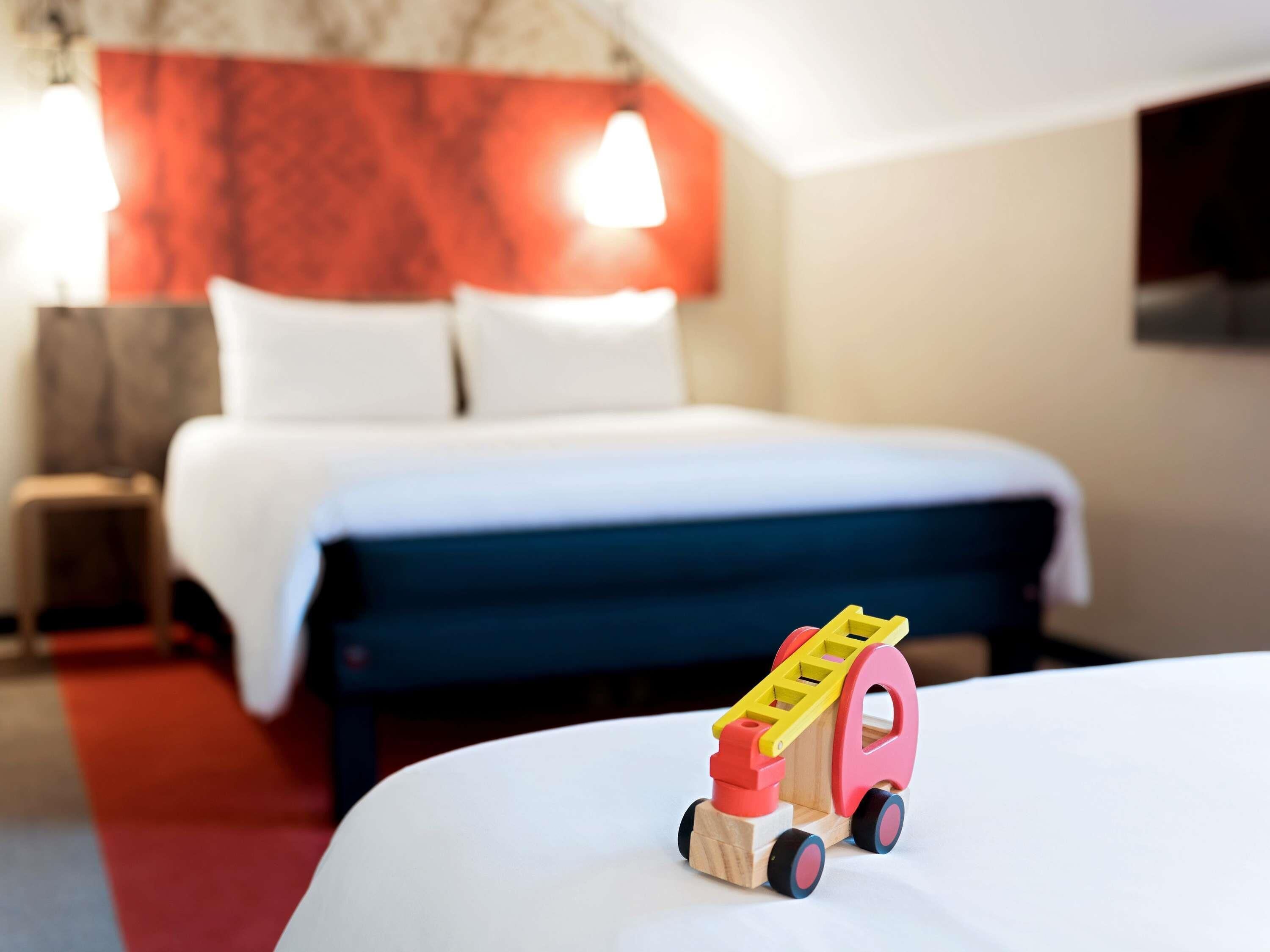 Quarto de hotel com uma cama, um pequeno caminhão de brinquedo de madeira e uma parede vermelha.