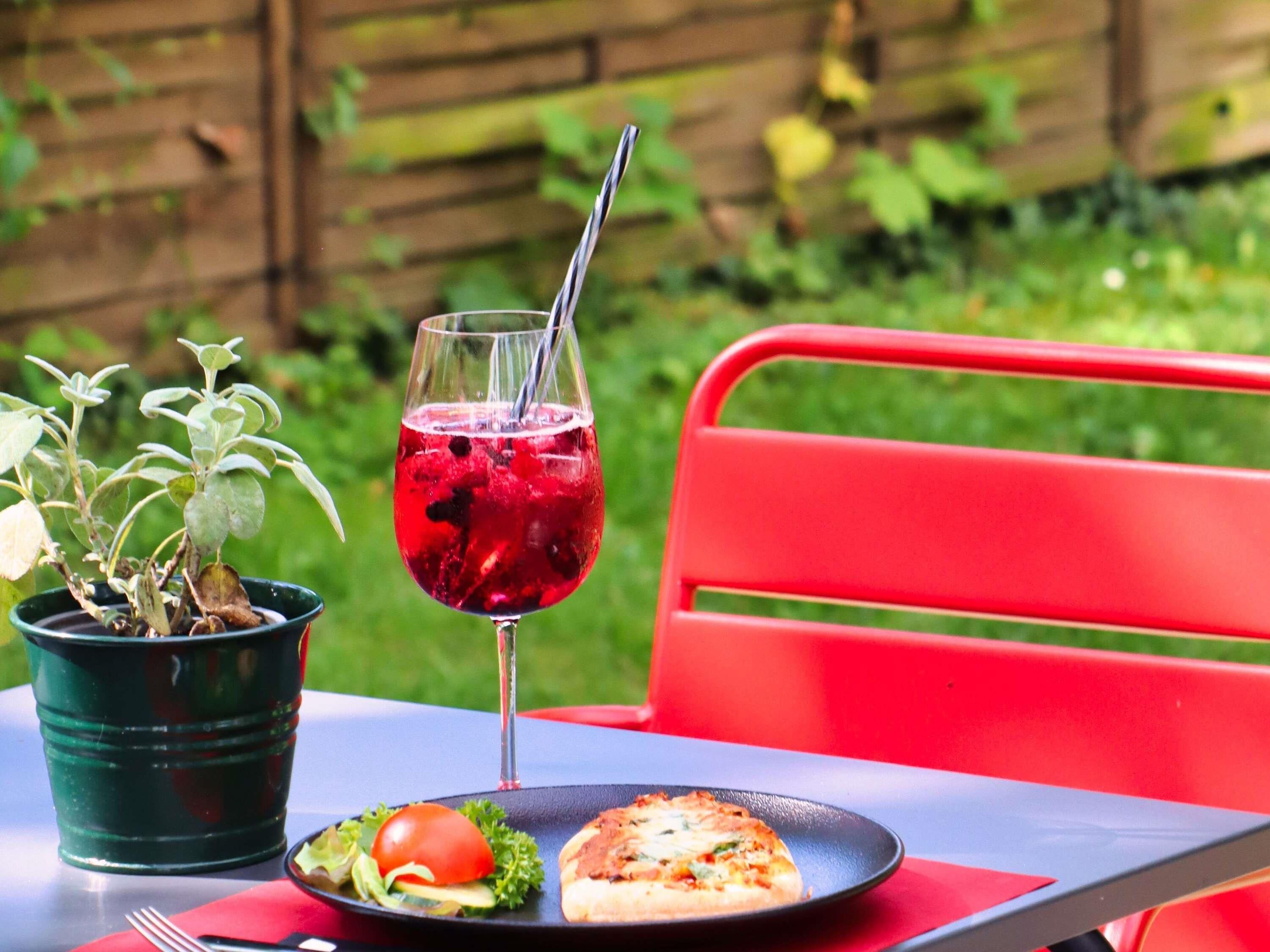A table with a glass of red drink, a plate with a sandwich and a tomato, and a potted plant.