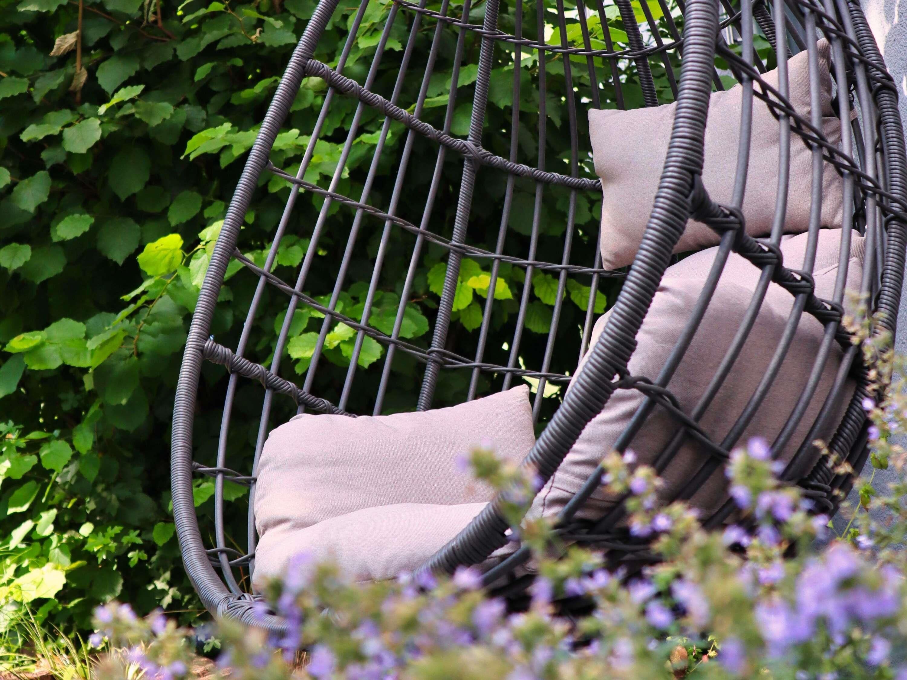 A wicker chair with cushions, surrounded by greenery and flowers.