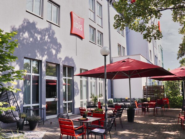 An outdoor seating area with tables and chairs, a red umbrella, and a building with a 'ibis' sign. - ibis Regensburg City (Regensburg)