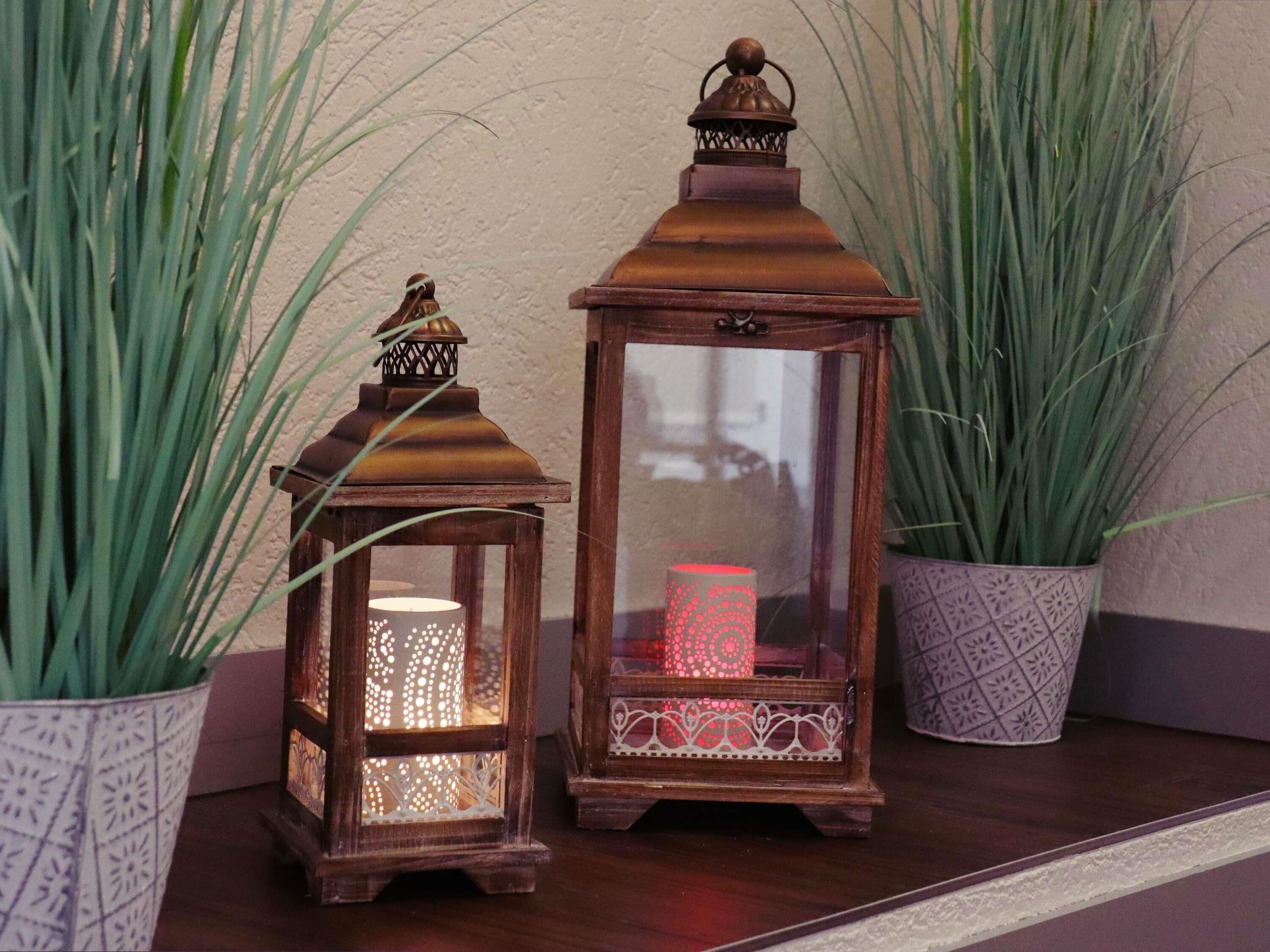 Two lanterns with intricate designs, placed on a wooden surface with tall green plants in patterned pots.