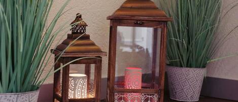 Two lanterns with intricate designs, placed on a wooden surface with tall green plants in patterned pots.