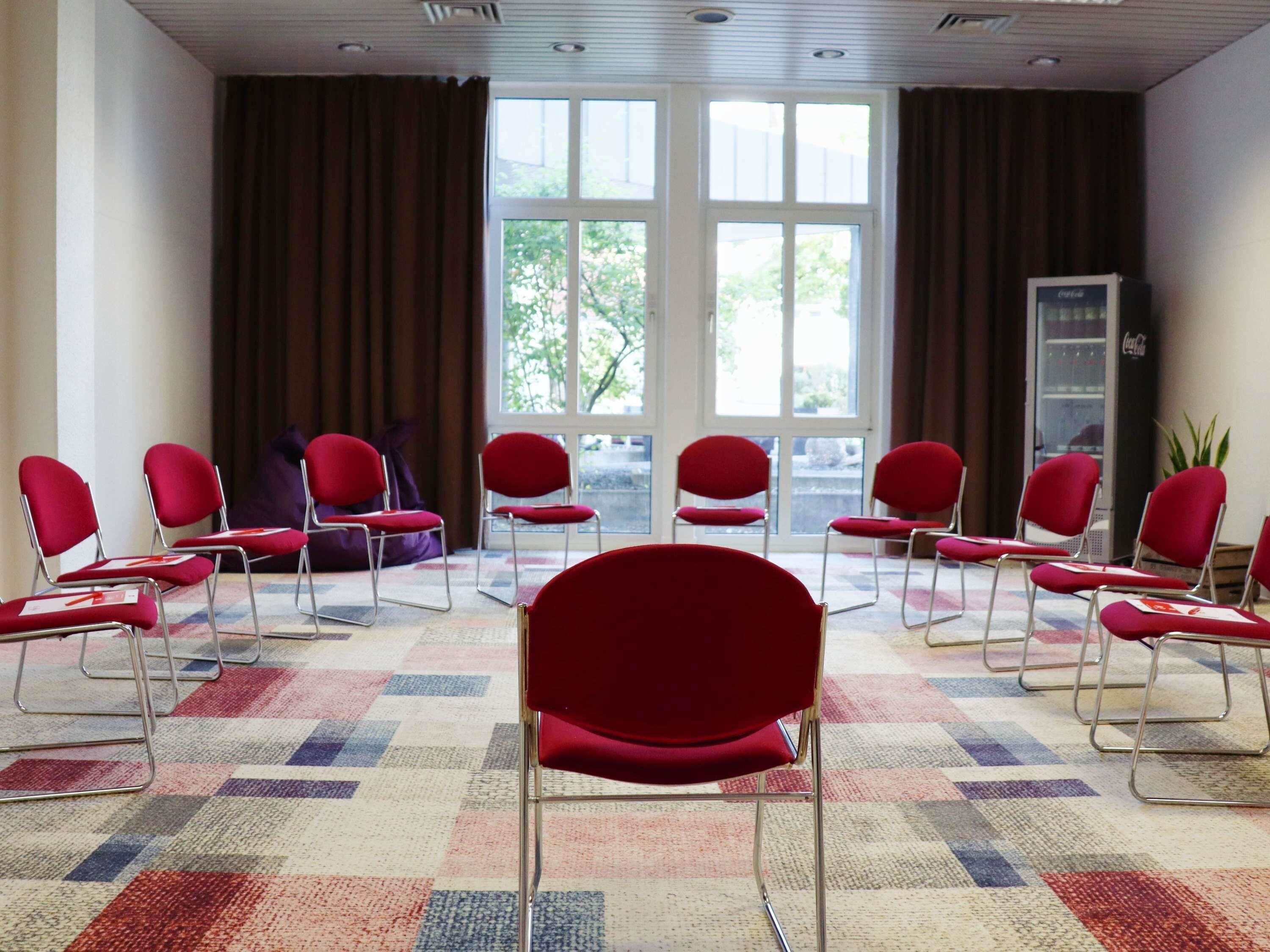 Un salón de conferencias con sillas rojas dispuestas en semicírculo, ventanas amplias con cortinas y una alfombra estampada.