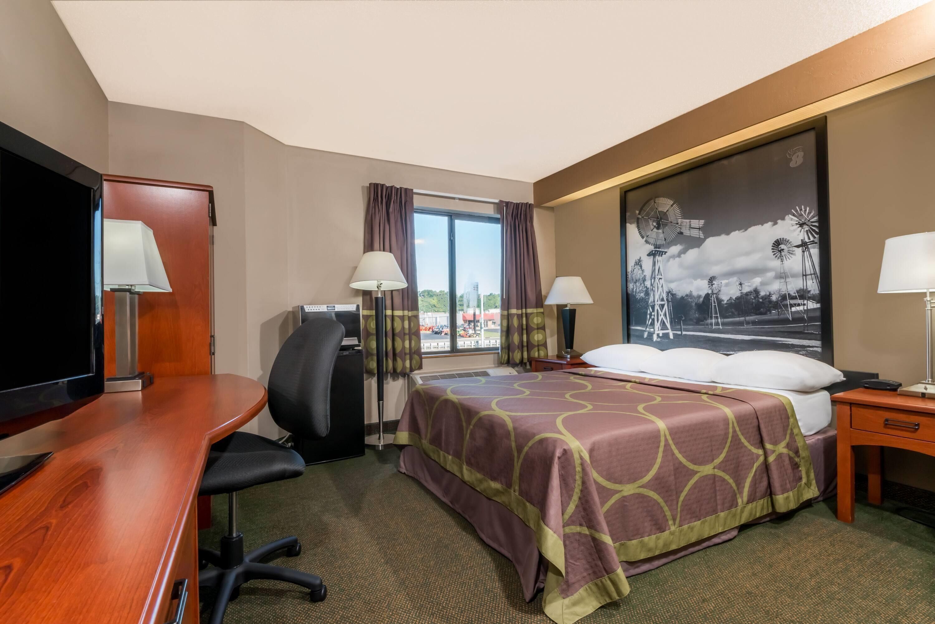 Habitación de hotel con cama, escritorio, silla, televisor y un amplio ventanal con cortinas.