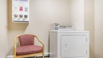 A laundry area with a chair, a washing machine, and a wall-mounted dispenser for bleach softeners.