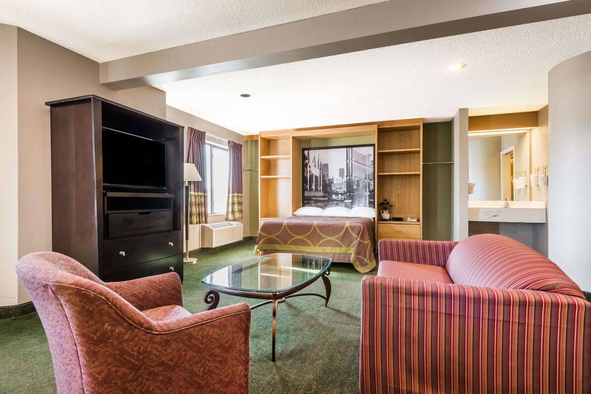 A hotel room with a bed, two armchairs, a glass coffee table, a TV, a bookshelf, and a large window.
