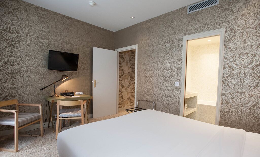Una habitación de hotel con cama, escritorio con silla, televisor montado en la pared y puerta que da a un baño.