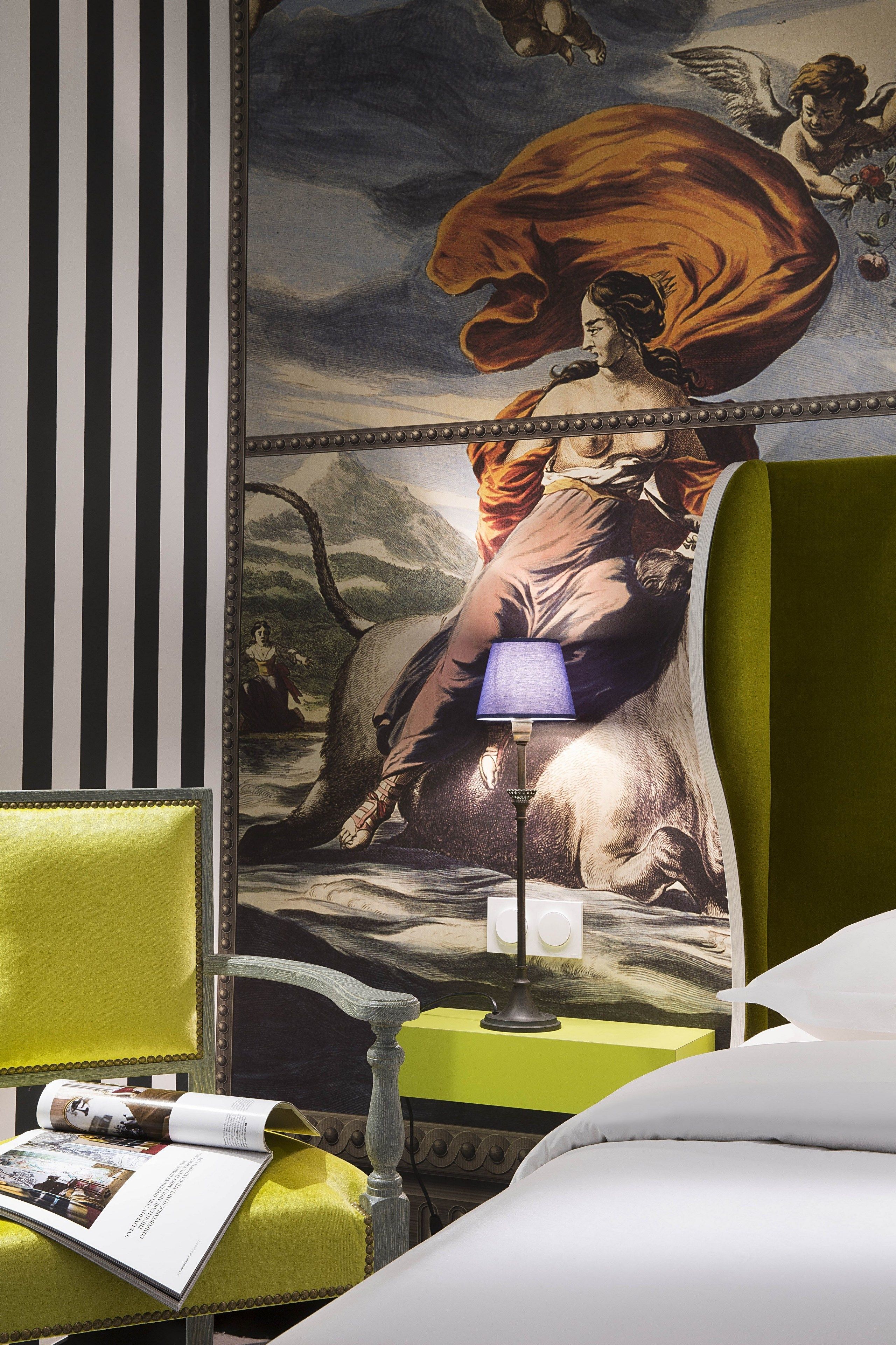 Une chambre d’hôtel avec une grande fresque murale, un fauteuil vert, une table de chevet avec une lampe et un lit avec du linge de lit blanc.