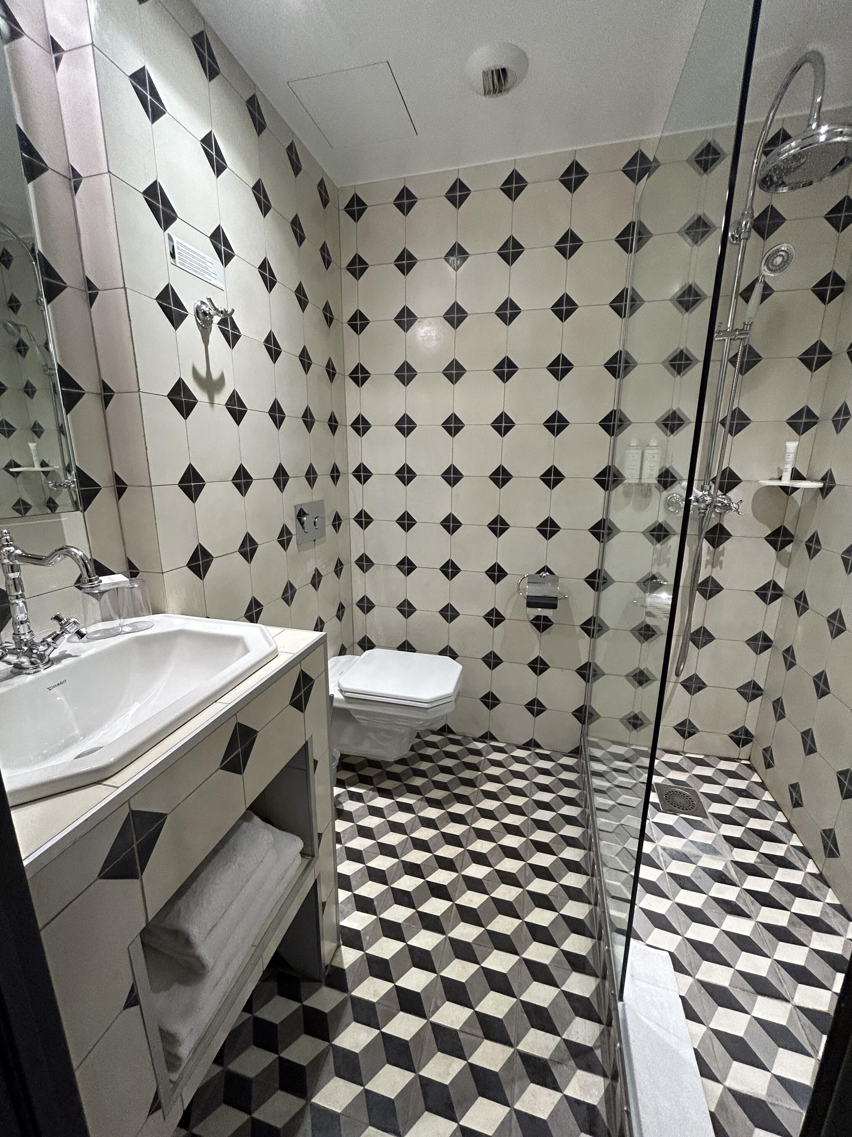 Un baño con piso de diseño geométrico, un lavamanos blanco, un inodoro y una zona de ducha con mampara de vidrio.