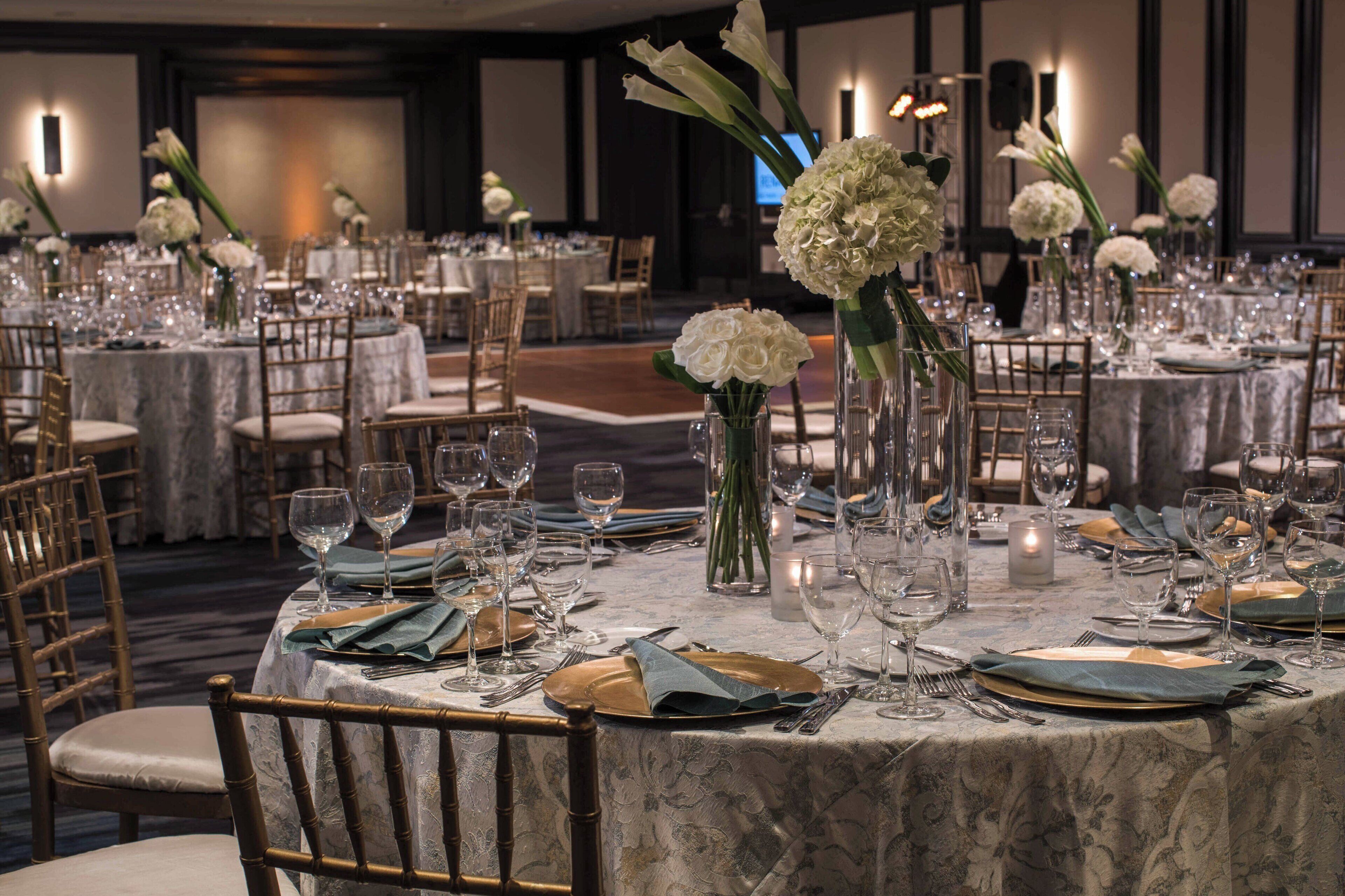 Un salón de banquetes con mesas redondas dispuestas para un evento formal, con sillas doradas, manteles blancos y centros de mesa florales.