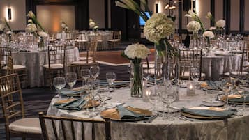 Un salón de banquetes con mesas redondas dispuestas para un evento formal, con sillas doradas, manteles blancos y centros de mesa florales.