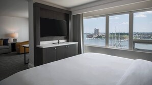A hotel room with a large bed, a flat-screen TV mounted on the wall, a small table with a lamp, and a view of a cityscape and water through large windows.