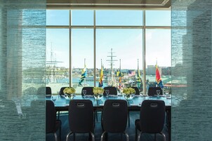 Un salón de conferencias con un amplio ventanal que da a un puerto con barcos y banderas.