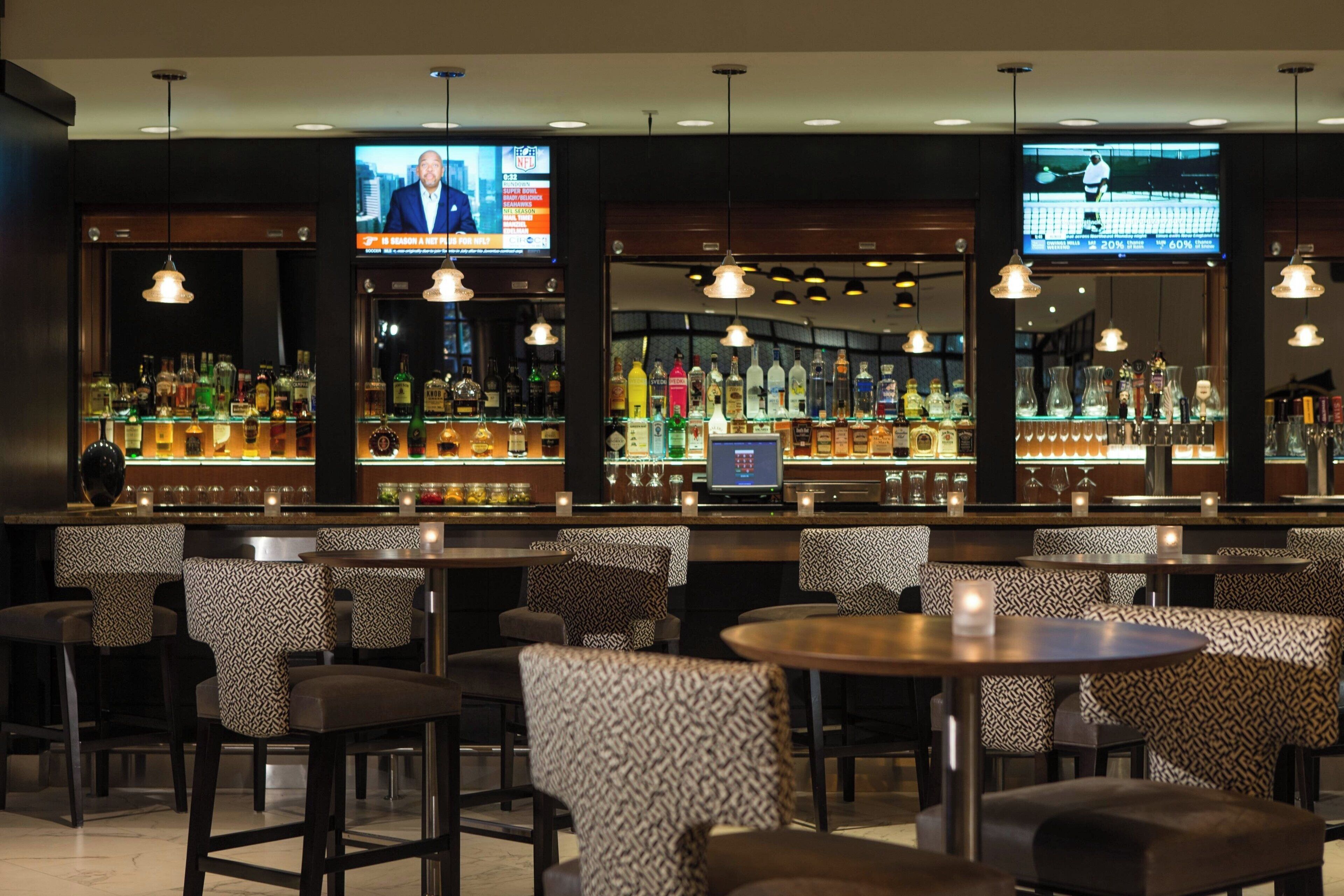 A bar area with multiple seating options, including bar stools and round tables. There are televisions mounted on the walls displaying news channels.