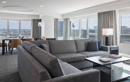 A modern hotel room with a large gray sectional sofa, a glass coffee table, and a view of the city through large windows.