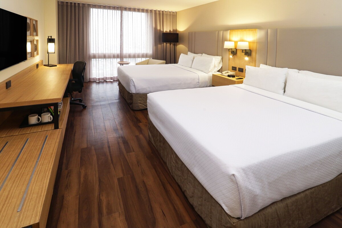A hotel room with two beds, a desk with a chair, a TV, and a large window with curtains.