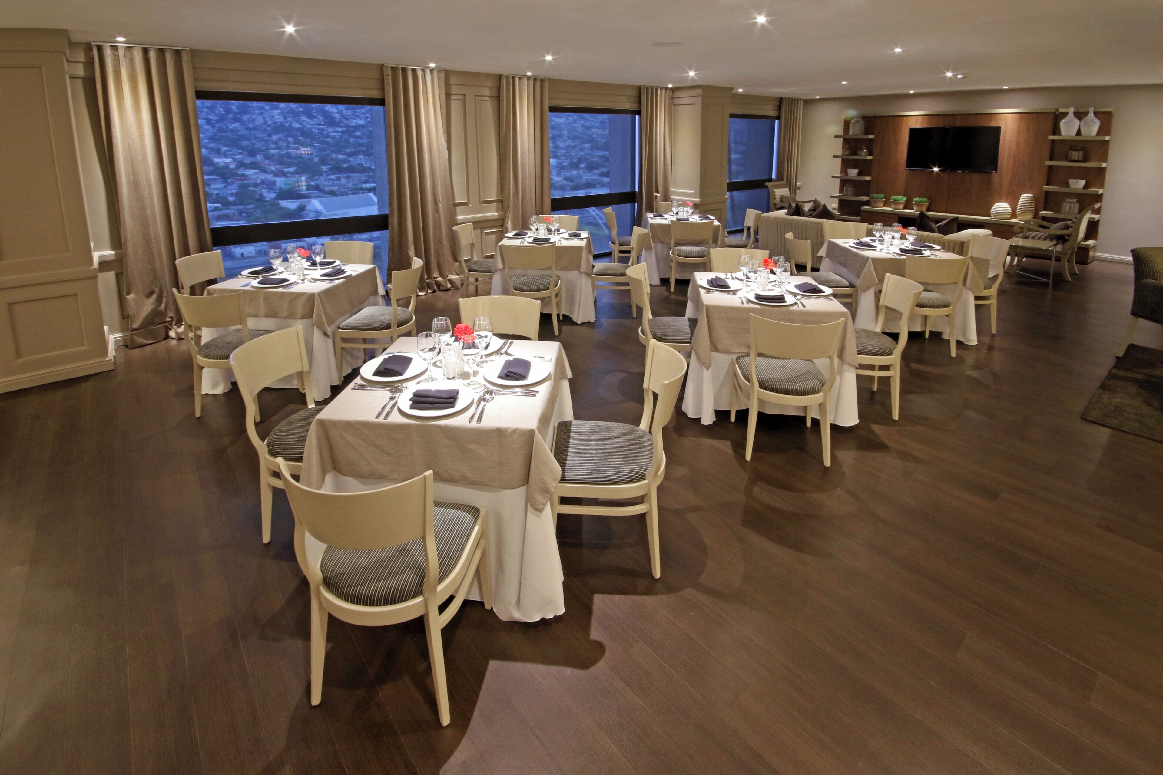 A dining area with multiple tables set for guests, large windows with curtains, and a television mounted on the wall.