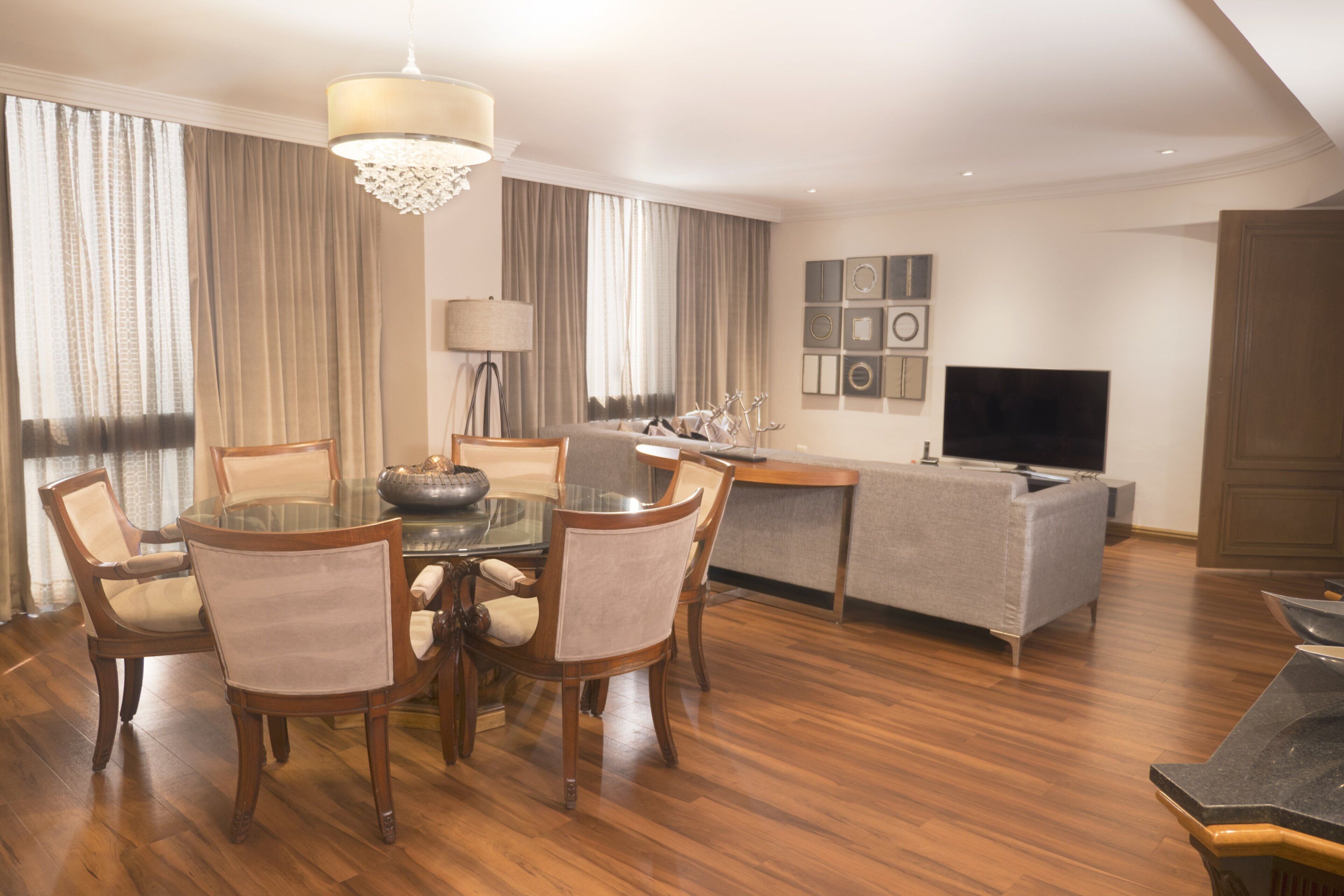 Una sala de estar moderna con comedor, que presenta una mesa redonda de vidrio, sillas de madera, un sofá gris, un televisor de pantalla plana y una araña.