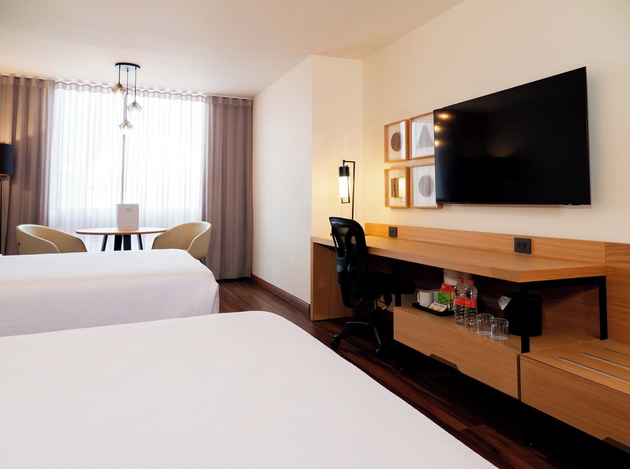 Ein Hotelzimmer mit zwei Betten, einem Schreibtisch mit Stuhl, einem Flachbildfernseher und einem Fenster mit Vorhängen.