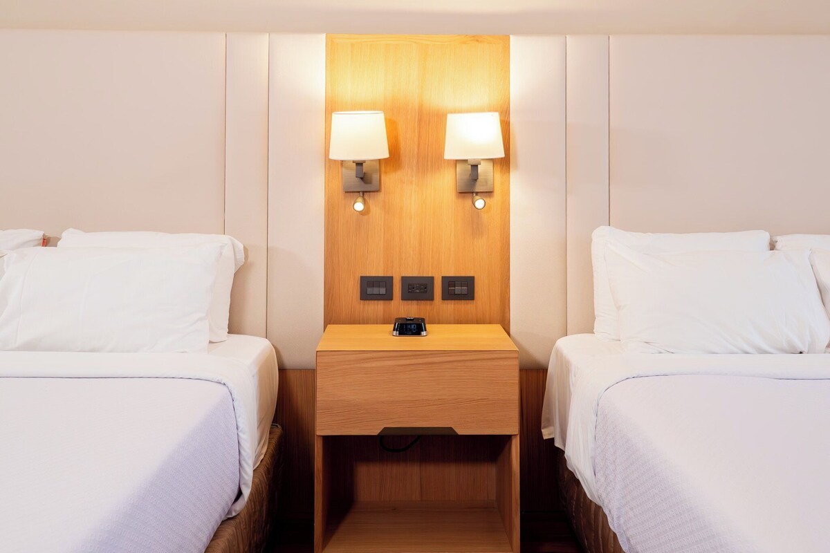 A hotel room with two beds, a wooden nightstand with two wall-mounted lamps, and a control panel.