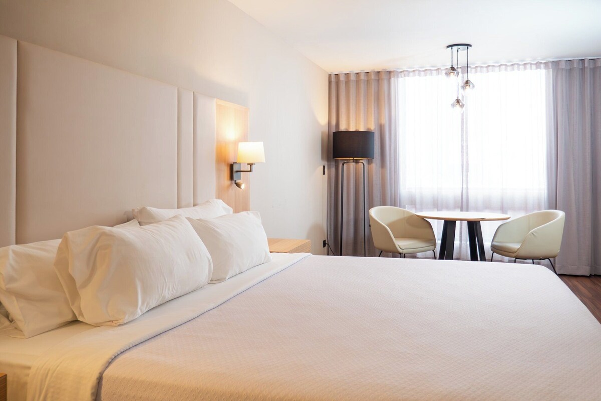 A hotel room with a large bed, two chairs, a table, a lamp, and a window with curtains.