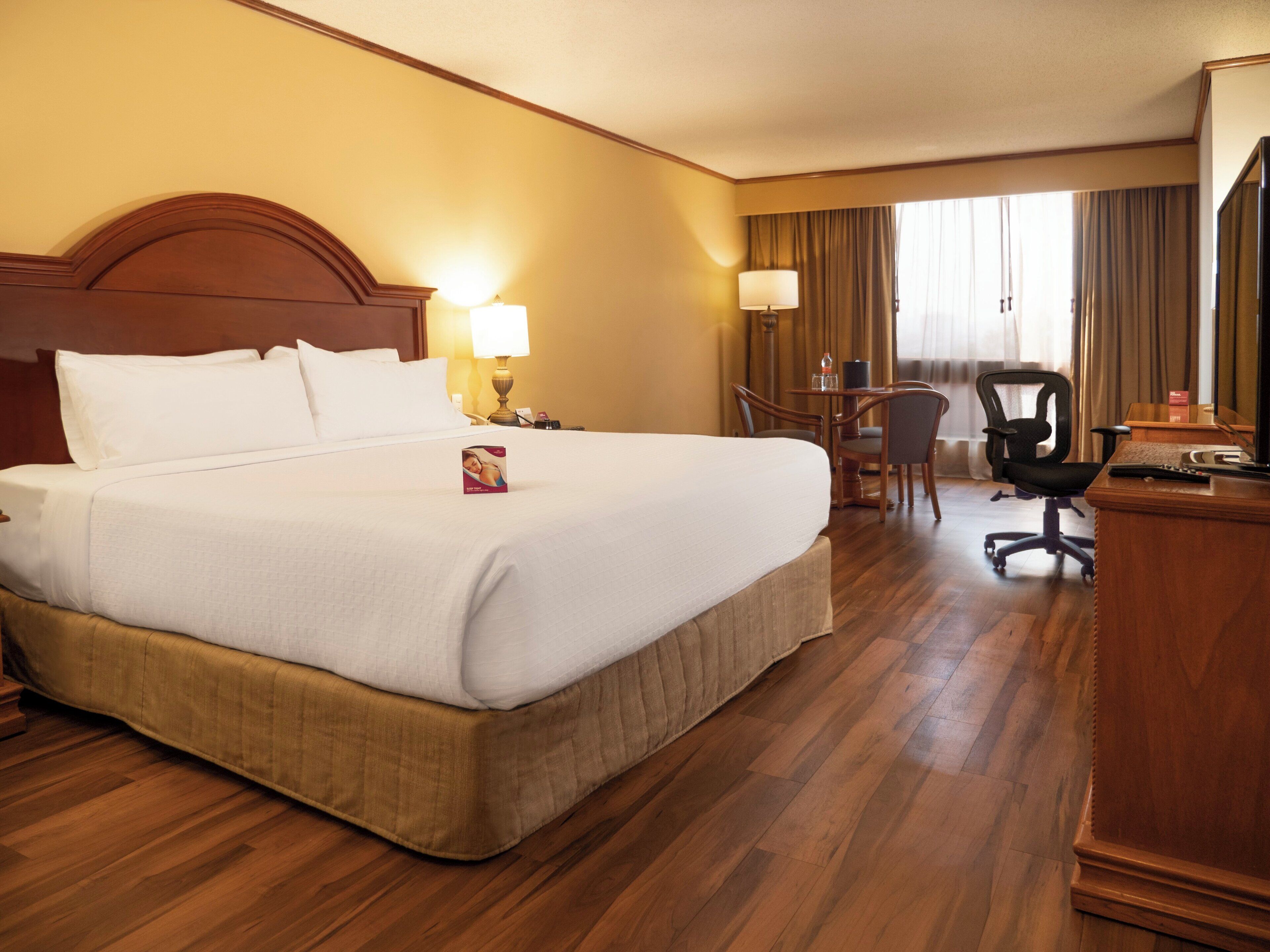 Habitación de hotel con una cama grande, un escritorio con silla, una lámpara y un ventanal con cortinas.
