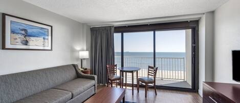 A hotel room with a sofa, a small table, two chairs, a TV, and a balcony with a view of the ocean.