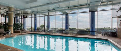 Une piscine intérieure avec des parois en verre transparent, un sol carrelé et une vue sur le ciel et les nuages.