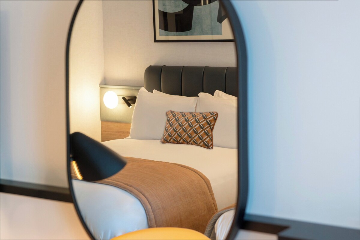 A hotel room with a bed, pillows, a nightstand, and a lamp.