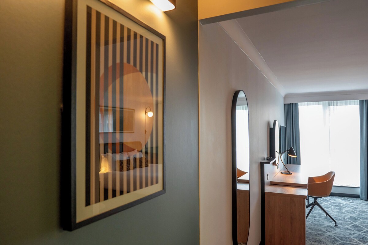 A hotel room with a framed artwork on the wall, a desk with a chair, a large mirror, and a window with curtains.