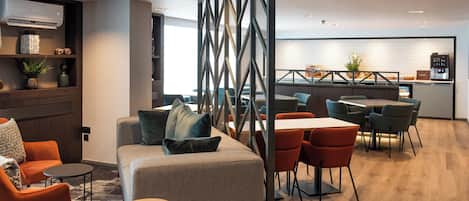 A modern hotel lobby with a seating area, a kitchen in the background, and a view of the city.