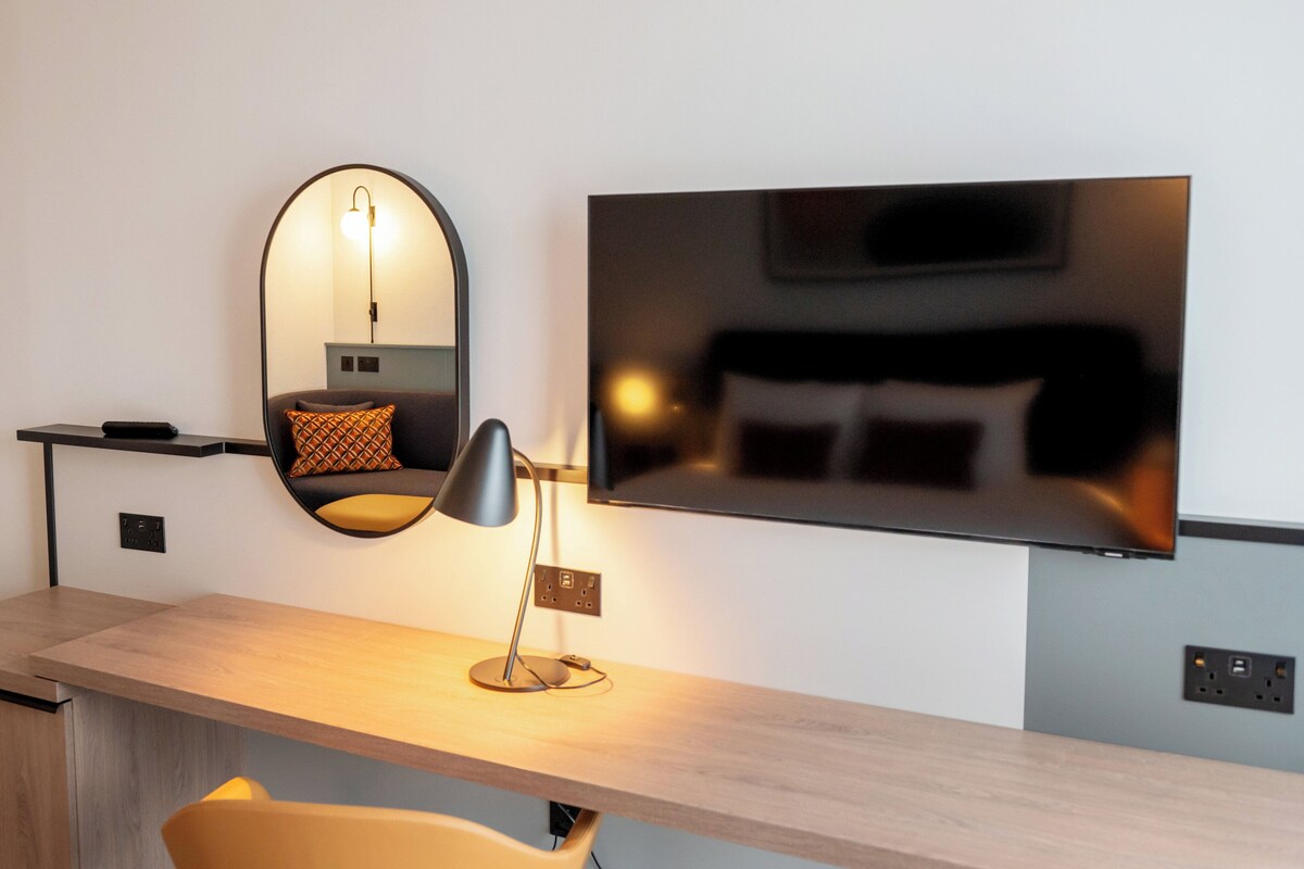 A modern hotel room with a desk, a large mirror, a flat-screen TV, and a bed with pillows.