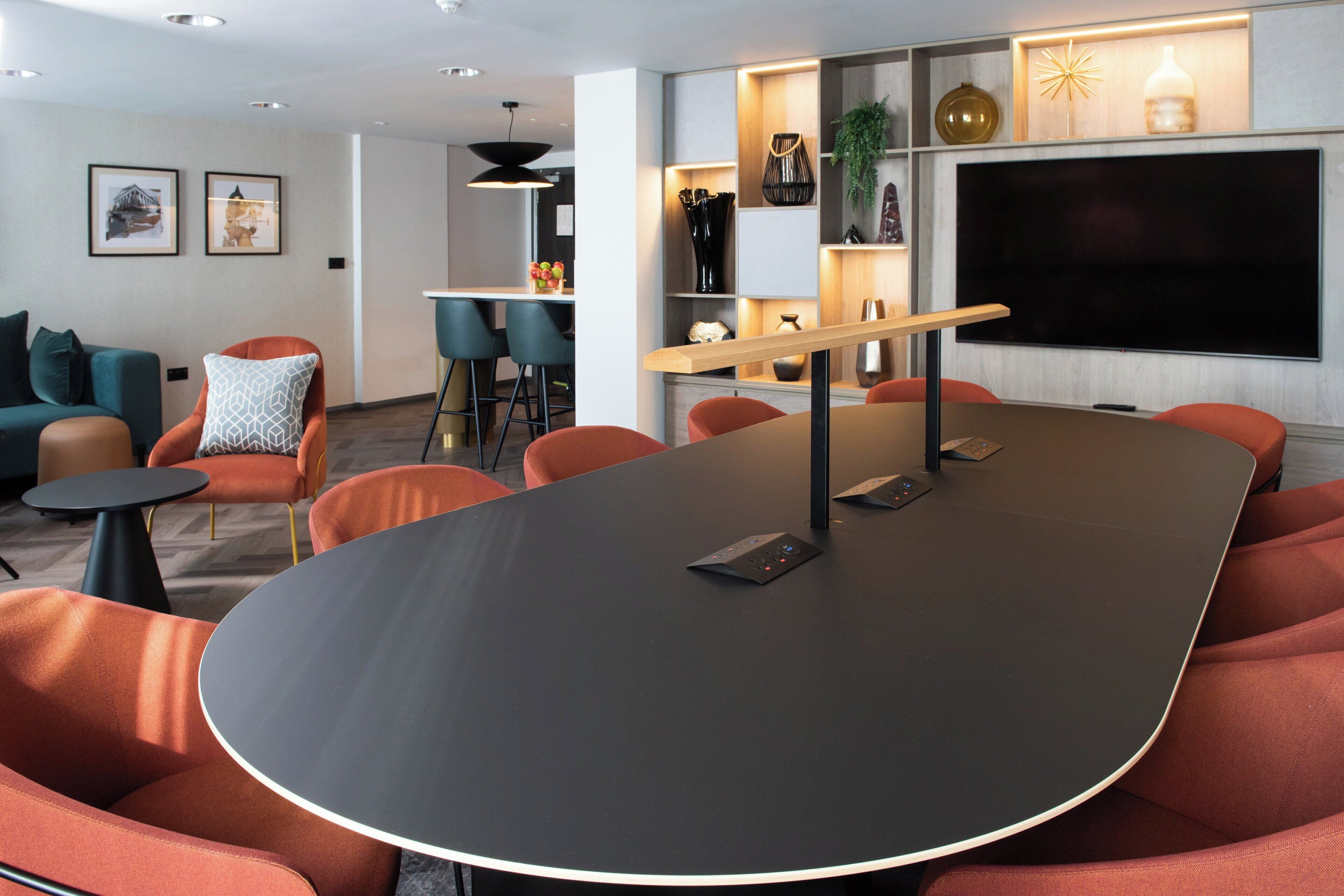 Una sala de conferencias moderna con una gran mesa, sillas color naranja, una zona de bar y un televisor montado en la pared.
