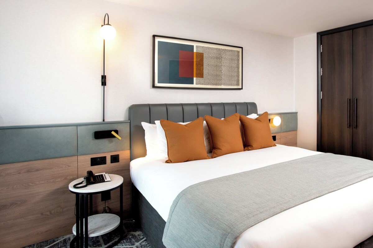 A hotel room with a bed, bedside table, lamp, artwork, wardrobe, and a small round table.