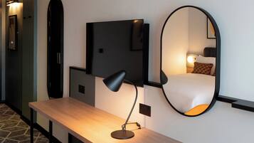 A hotel room with a wooden desk, a black desk lamp, a wall-mounted TV, and a large mirror reflecting a bed with a pillow.