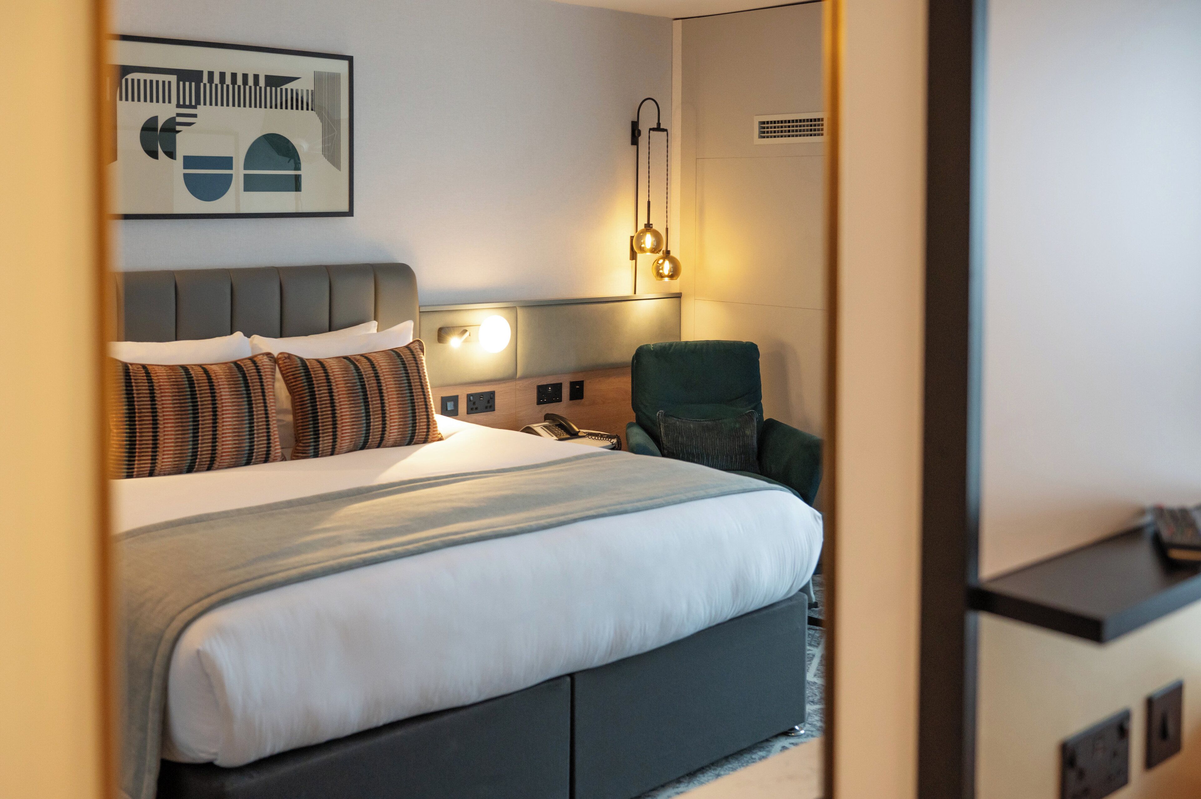 A hotel room with a bed, a green armchair, a wall-mounted phone, and a wall art piece.
