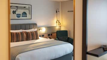 A hotel room with a bed, a green armchair, a wall-mounted phone, and a wall art piece.